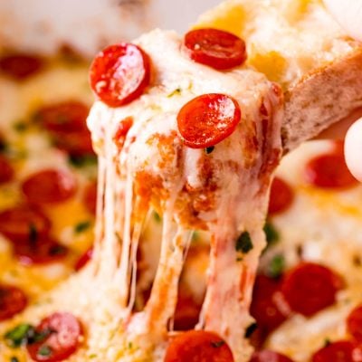 A piece of baguette scooping pizza dip our of a white baking dish.