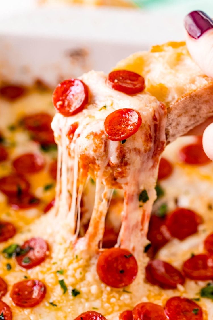 A piece of baguette scooping pizza dip our of a white baking dish.