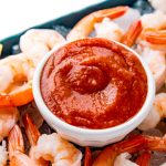 Close up photo of cocktail sauce in a small white bowl on top of a platter of shrimp and ice.