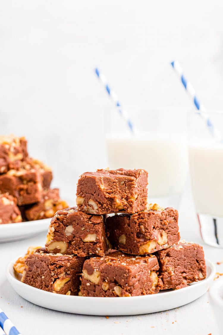 Walnut chocolate fudge stacked on a white plate with glasses of milk in the background.