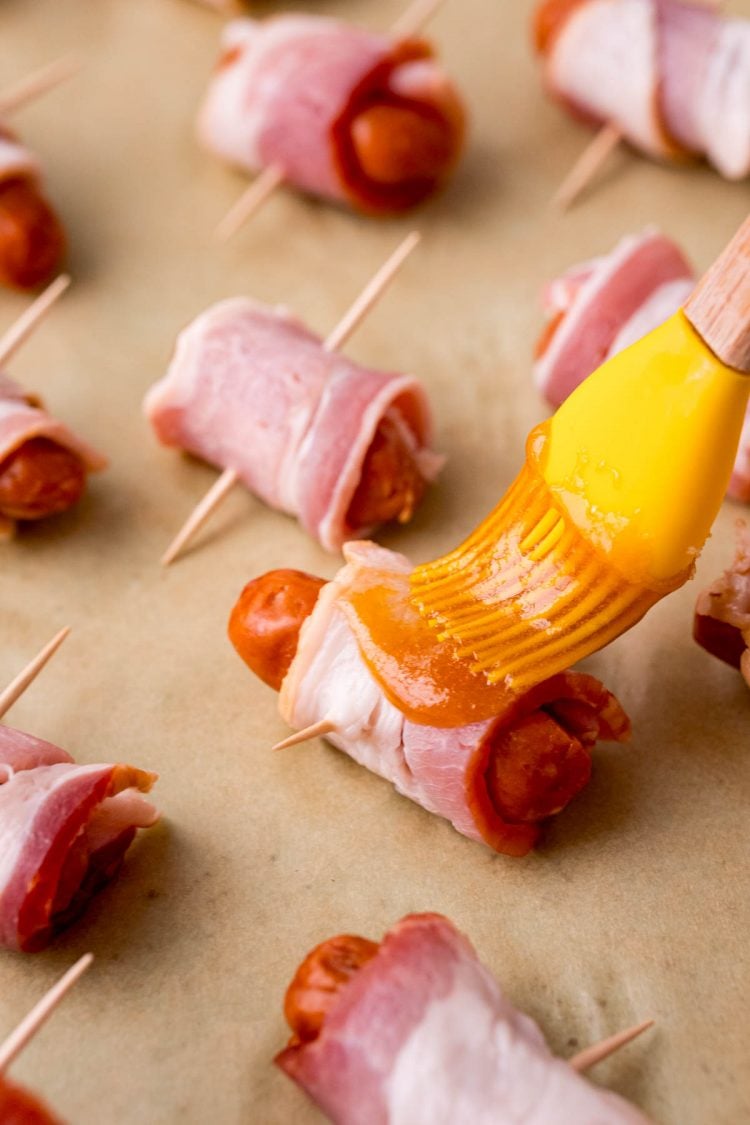 Close up photo of bacon wrapped little smokies on parchment paper being brushed with sauce.