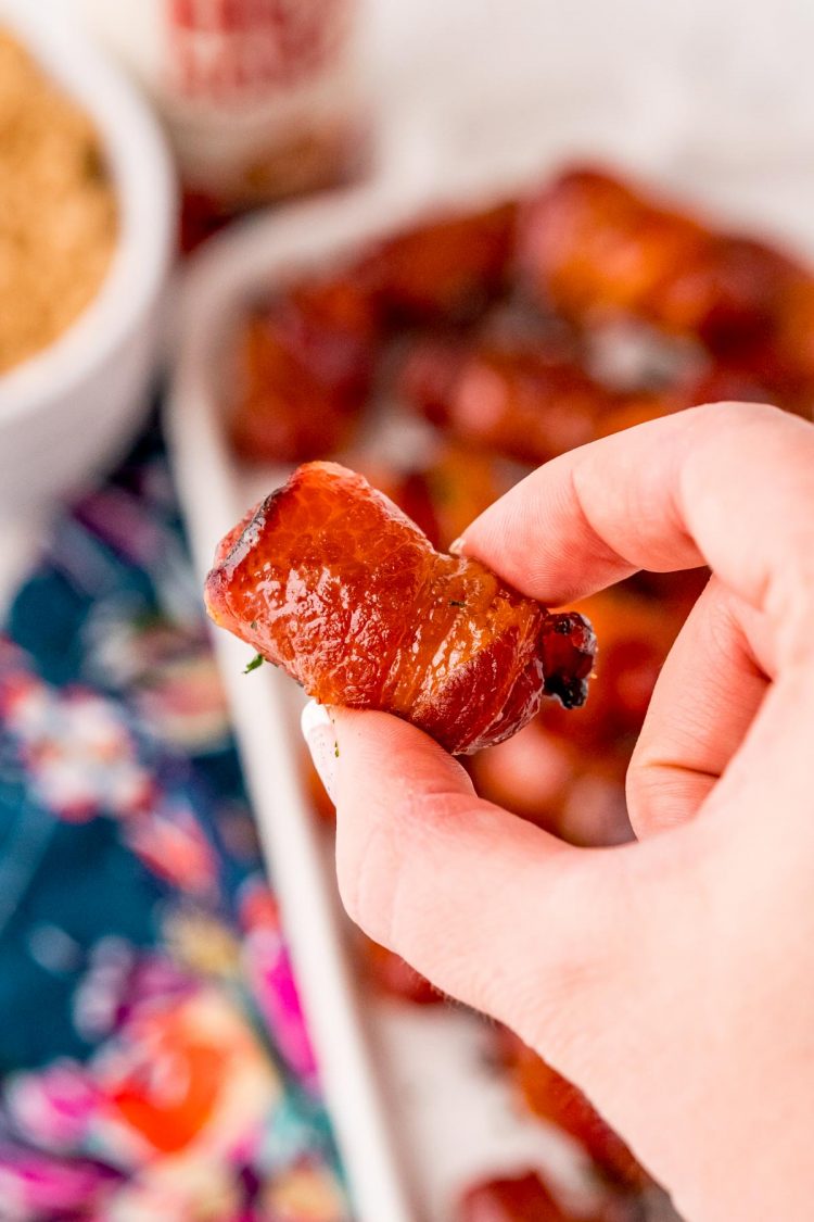 Close up photo of a hand holding a bacon wrapped little smokies. 