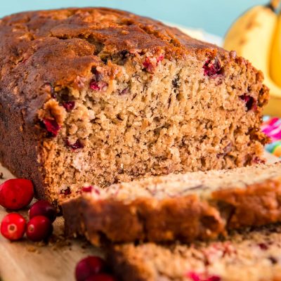 Close up photo of a loaf of cranberry banana bread that had been sliced halfway and is resting on a wooden cutting board.