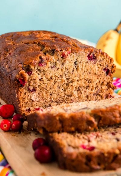 Close up photo of a loaf of cranberry banana bread that had been sliced halfway and is resting on a wooden cutting board.