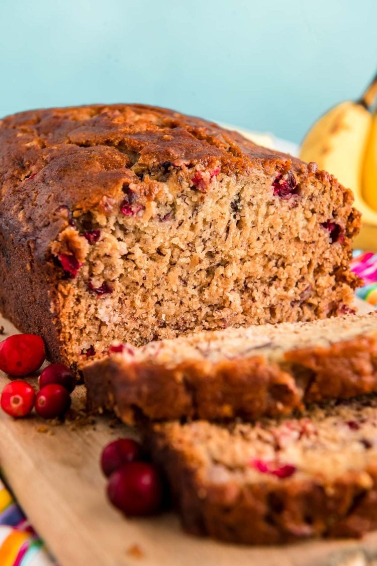 Close up photo of a loaf of cranberry banana bread that had been sliced halfway and is resting on a wooden cutting board.