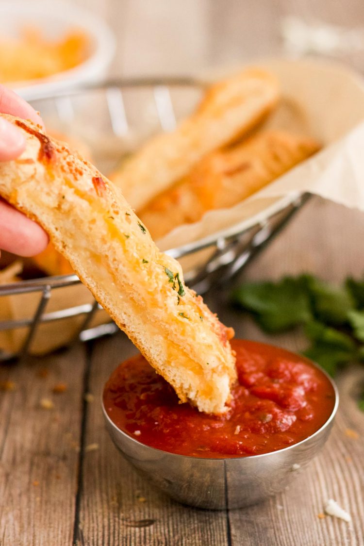 A breadsticks is being dipped in a small metal bowl of marinara sauce.