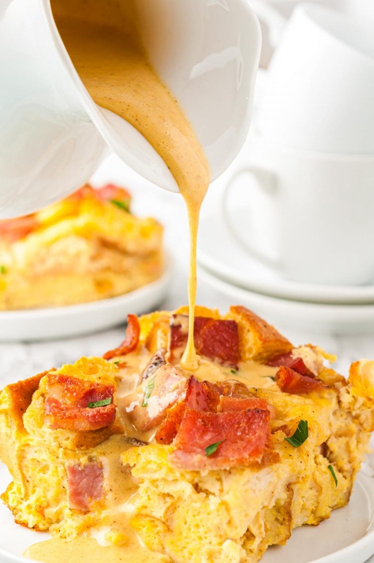 Hollandaise sauce being poured on a slice of eggs benedict breakfast casserole.