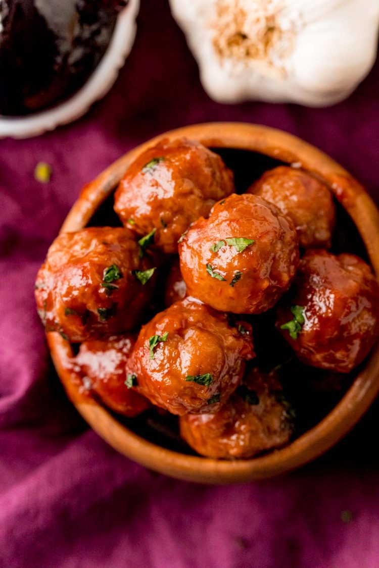 Close up photo of grape jelly meatballs in a wooden bowl on a purple napkin.