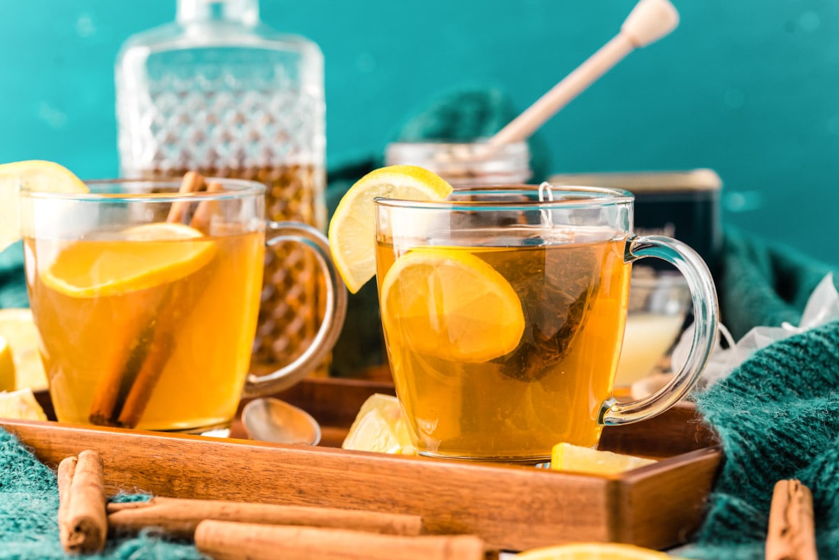 2 mugs on a wooden tray filling with tea, lemon, honey, and whiskey.