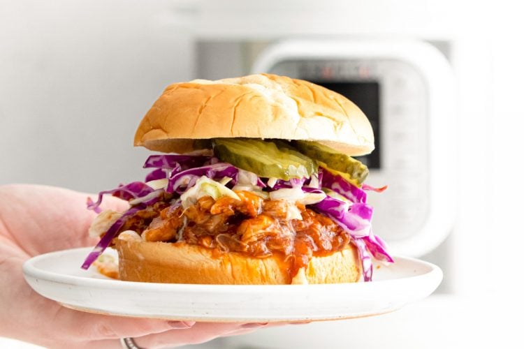 A woman's hand holding a white plate with a bbq chicken sandwich on it.