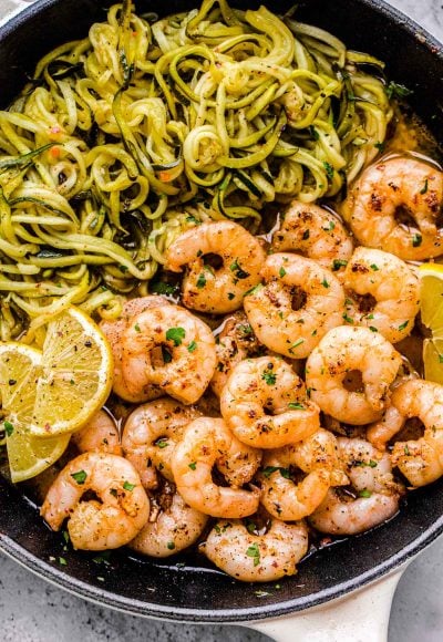 Close up photo of lemon garlic shrimp in a skillet with zucchini noodles and lemon wedges.