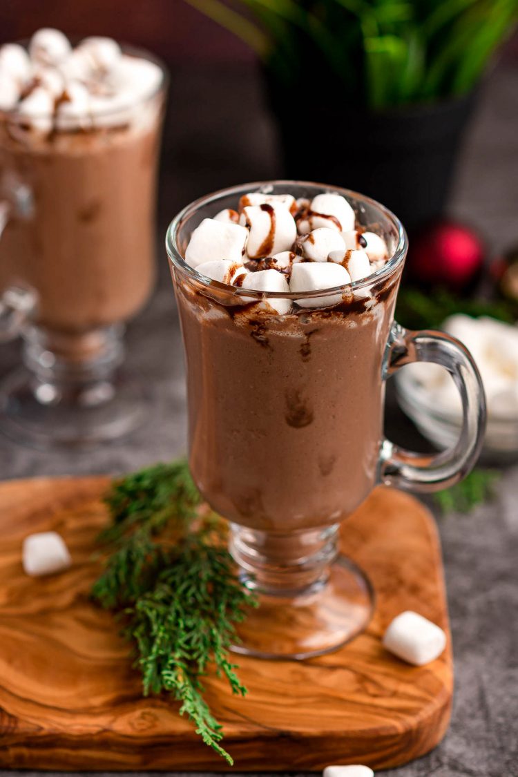 A glass mug of hot chocolate on a wooden cutting board.