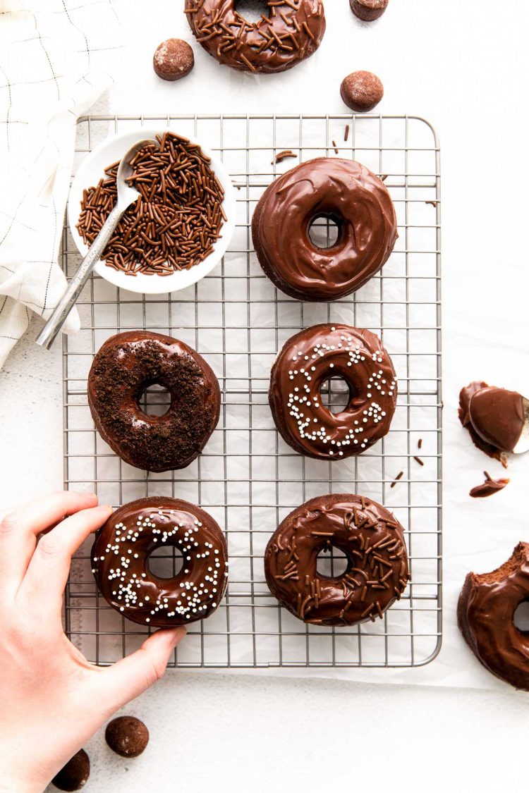 A woman's hand grabbing a chocolate frosted donut off of a wire rack.