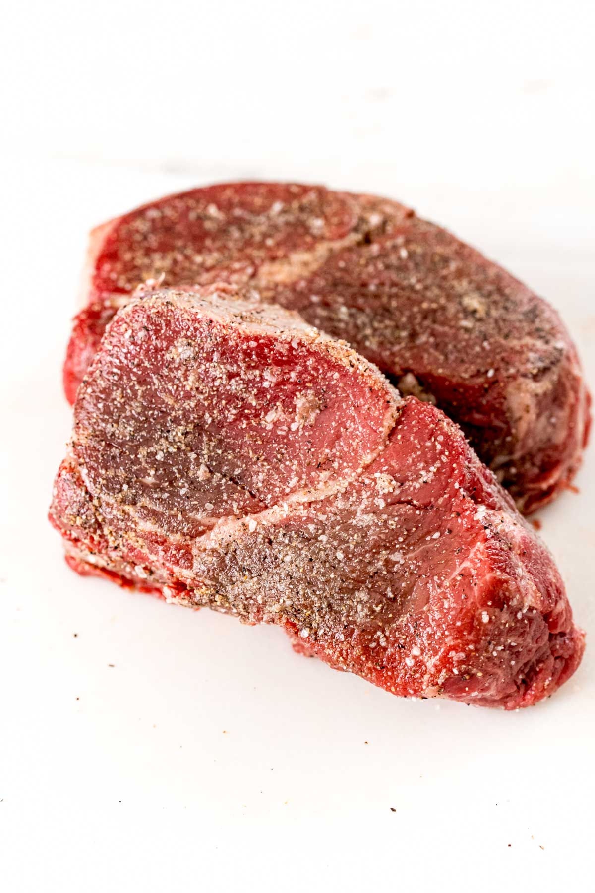 Raw filet mignon on a white cutting board seasoned with oil, salt, pepper, and garlic powder.