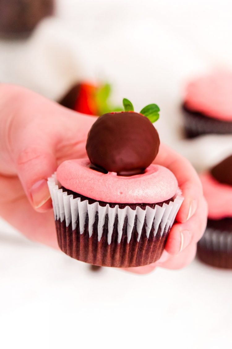 A woman's hand holding a chocolate covered strawberry cupcake.