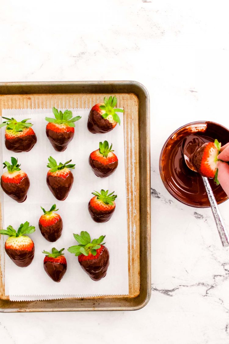 Chocolate covered strawberries on a baking sheet and a woman's hand dipping more in chocolate.