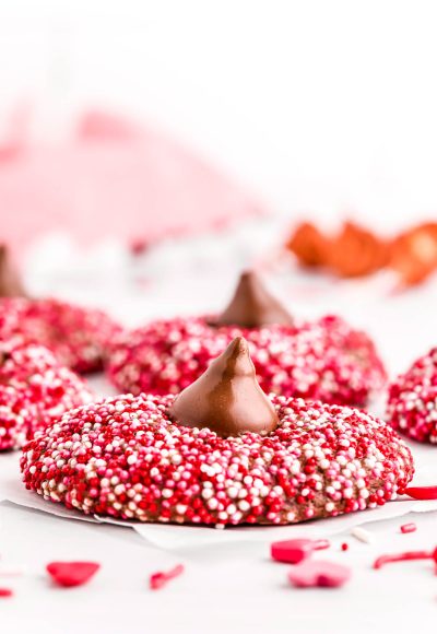 Close up photo of chocolate cookies covered in red, pink, and white sprinkles and topped with a hershey kiss.