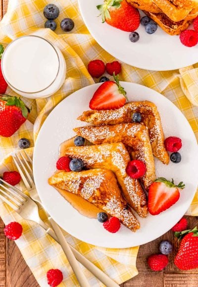Overhead photo of a plate of french toast that has been slices into triangles and topped with powdered sugar, syrup, and fruit on a yellow napkin with milk next to it.