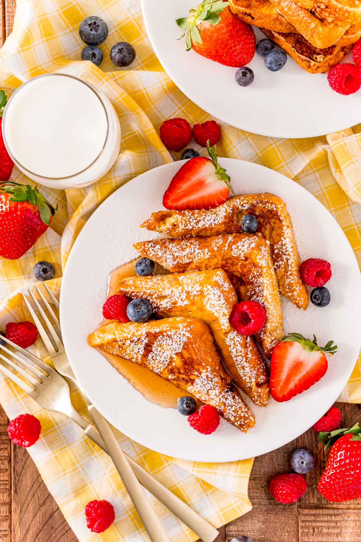 Overhead photo of a plate of french toast that has been slices into triangles and topped with powdered sugar, syrup, and fruit on a yellow napkin with milk next to it.