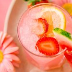 A close up photo of a glass with a pink cocktail in it garnished with strawberries and lemon.