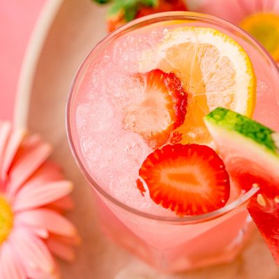 A close up photo of a glass with a pink cocktail in it garnished with strawberries and lemon.