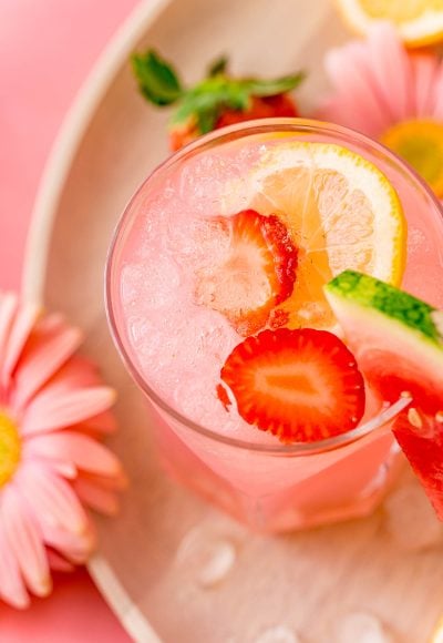 A close up photo of a glass with a pink cocktail in it garnished with strawberries and lemon.