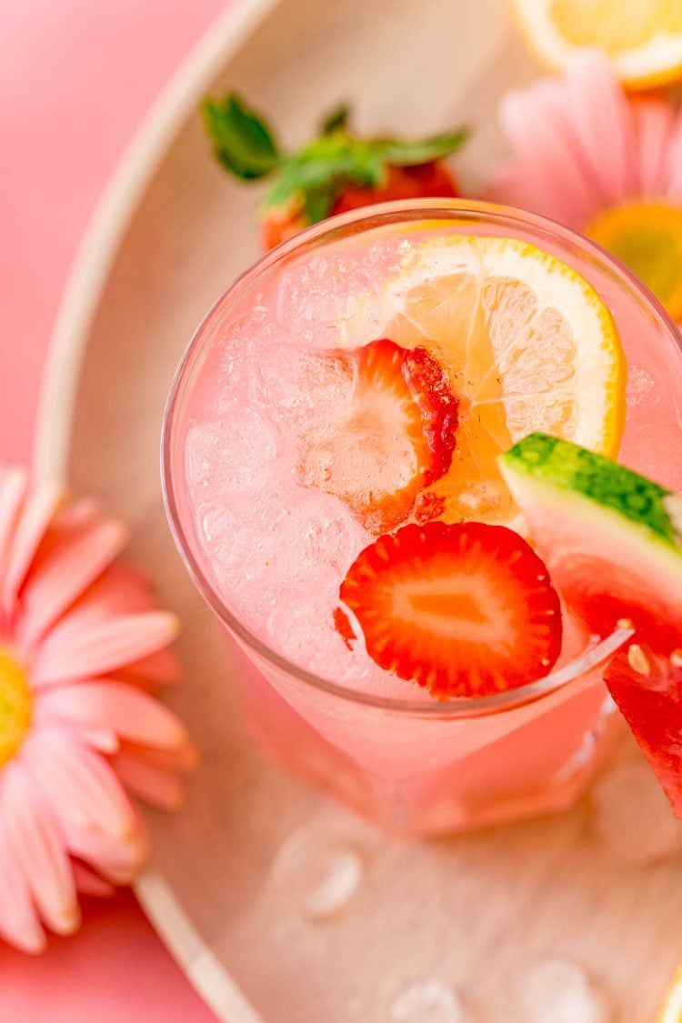A close up photo of a glass with a pink cocktail in it garnished with strawberries and lemon.