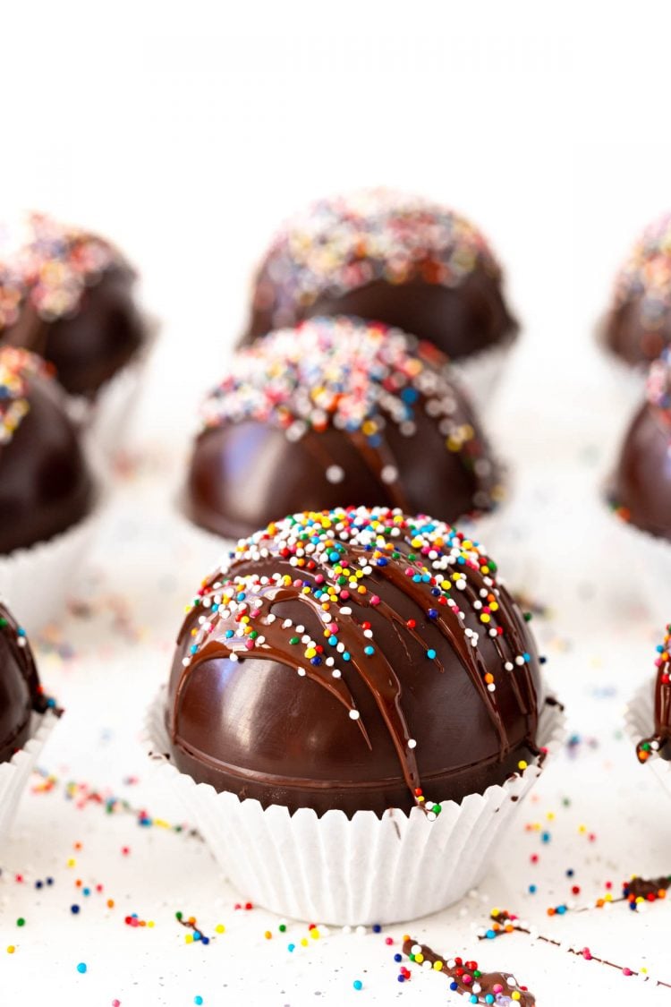 Close up photo of hot chocolate bombs topped with sprinkles on a white surface more more in the background.