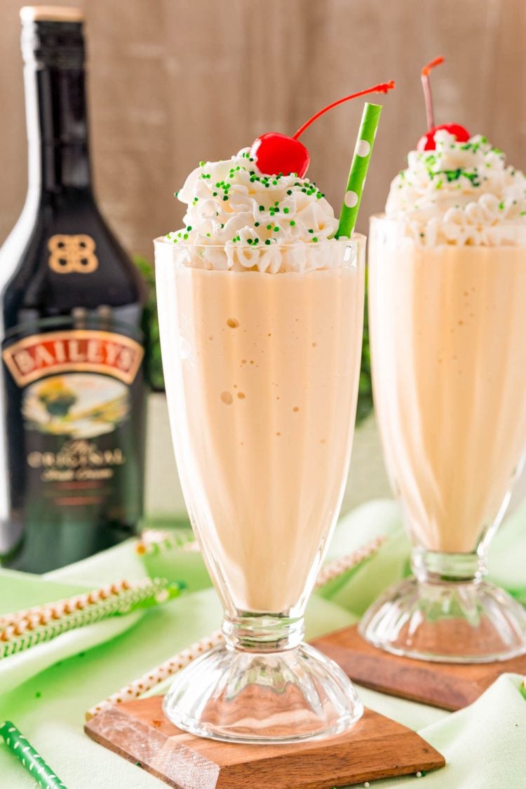 Two Irish cream milkshakes on wood coasters on a green napkin with a bottle of Bailey's in the background.