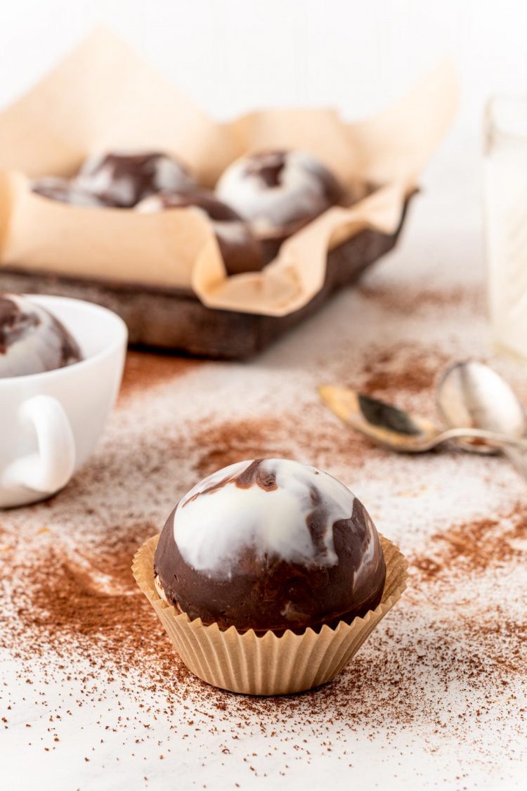Close up photo of a mocha hot chocolate bomb on a white table with cocoa powder around it and more cocoa bombs in the background.
