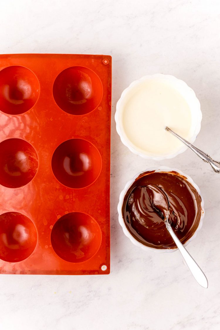 Overhead photo of hot chocolate bomb molds with bowls of melted white chocolate and dark chocolate next to it.