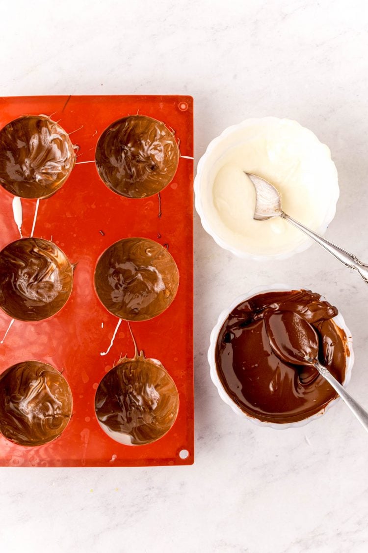 Hot chocolate bomb mold filled with chocolate with bowls of melted white chocolate and dark chocolate next to it.