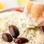 A piece of pita bread being dipped in a bowl of tzatziki sauce.