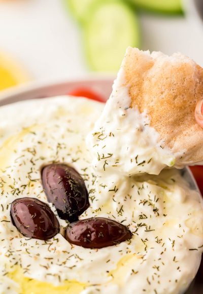 A piece of pita bread being dipped in a bowl of tzatziki sauce.