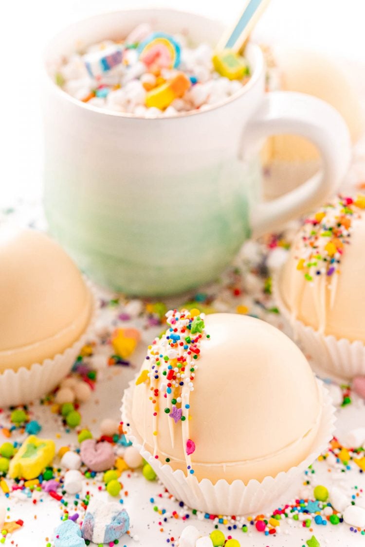 White hot cocoa bombs with a mug in the background surrounded by sprinkles and lucky charms marshmallows.