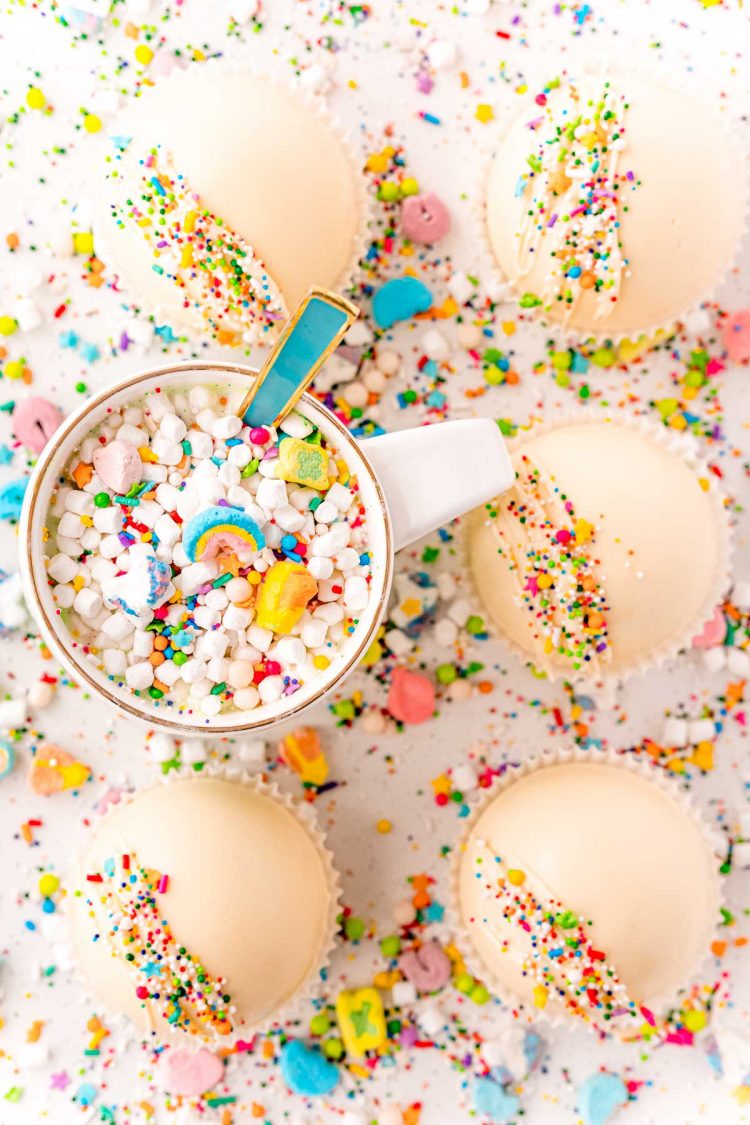 Overhead photo of white hot cocoa bombs on a white counter surrounded by sprinkles and a mug of cocoa.