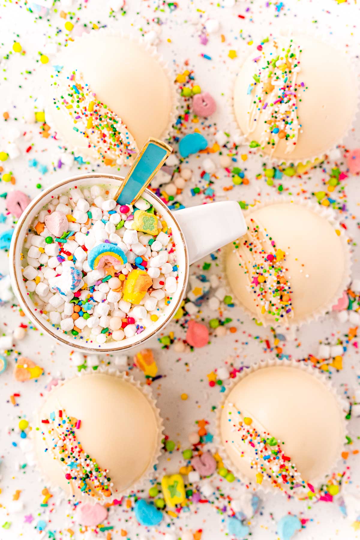 Overhead photo of white hot cocoa bombs on a white counter surrounded by sprinkles and a mug of cocoa.