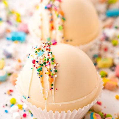 Close up photo of white chocolate hot chocolate bombs on a white table with rainbow sprinkles all around.