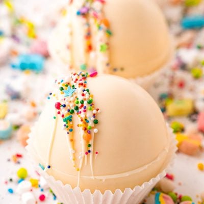 Close up photo of white chocolate hot chocolate bombs on a white table with rainbow sprinkles all around.