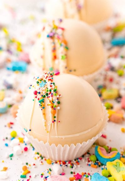 Close up photo of white chocolate hot chocolate bombs on a white table with rainbow sprinkles all around.