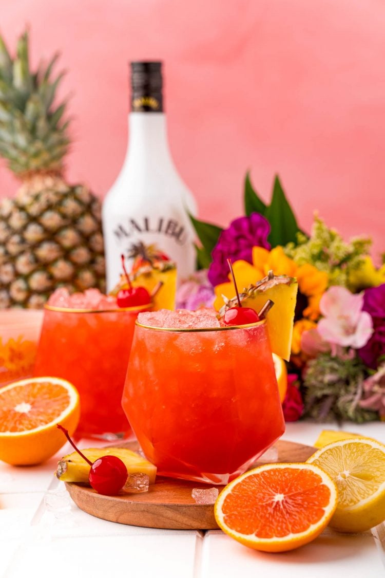 Two Bahama Mama cocktails on a wooden board on a white table with a bottle of rum in the background and oranges and pineapple around.