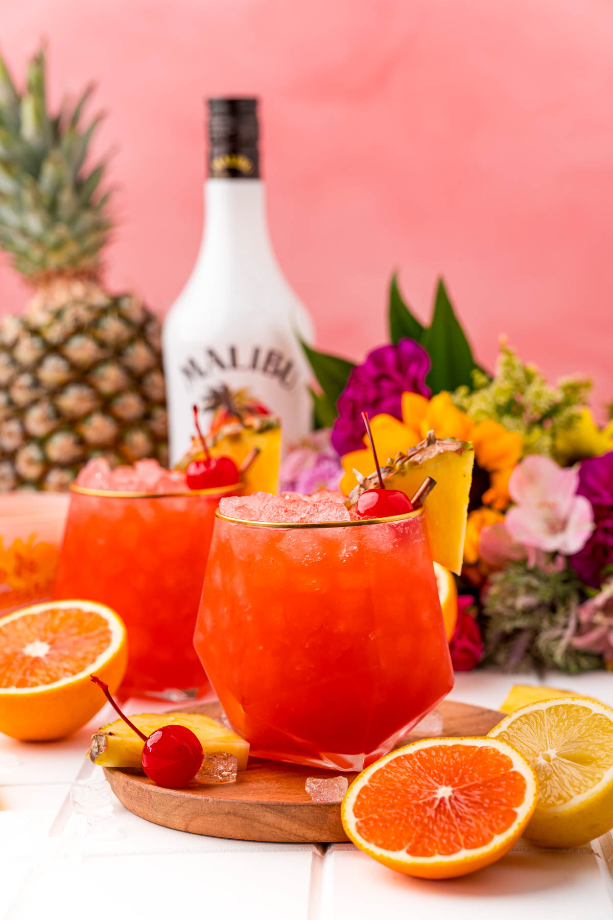 Two Bahama Mama cocktails on a wooden board on a white table with a bottle of rum in the background and oranges and pineapple around.