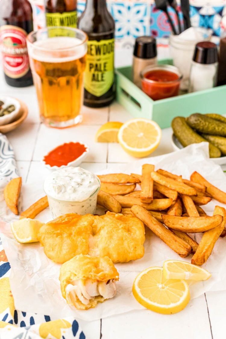 Fish and chips on a counter with beer and lemons and tartar sauce in the background.