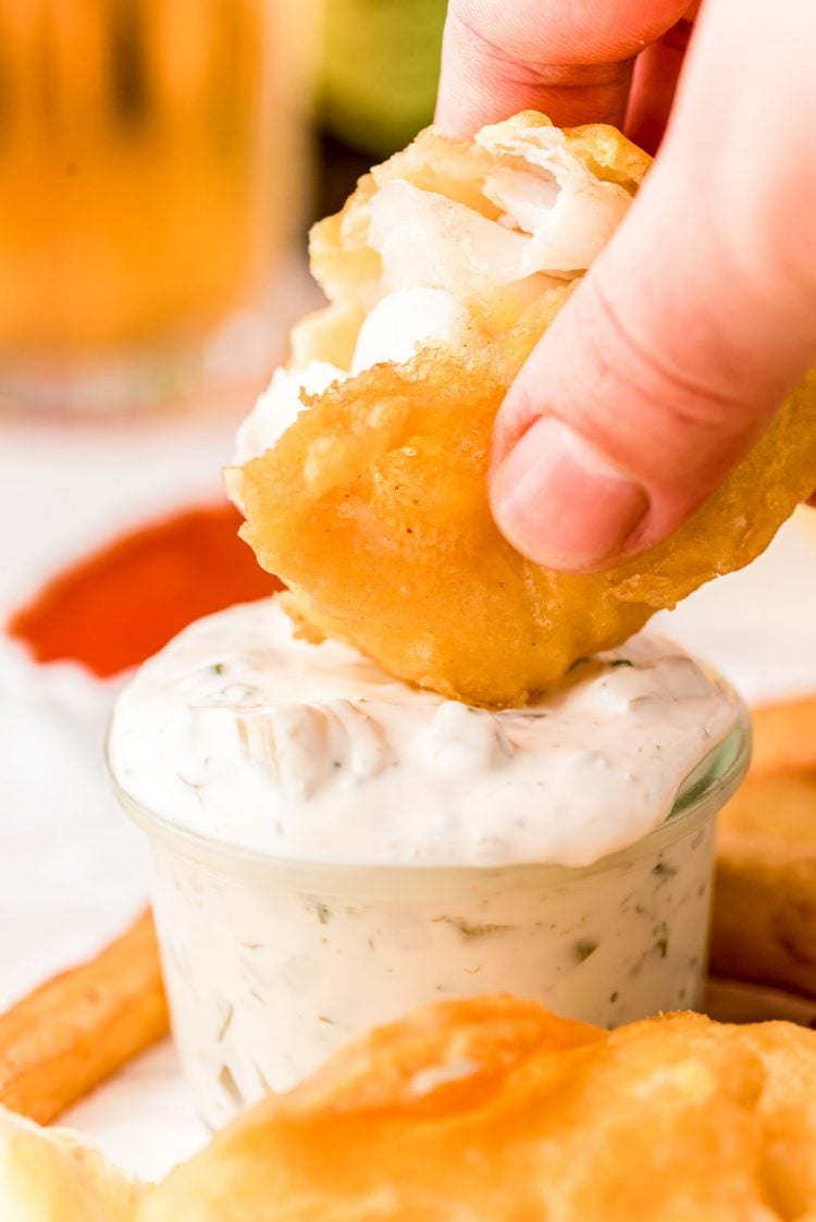 A woman's hand dipping a piece of fried fish in tartar sauce.