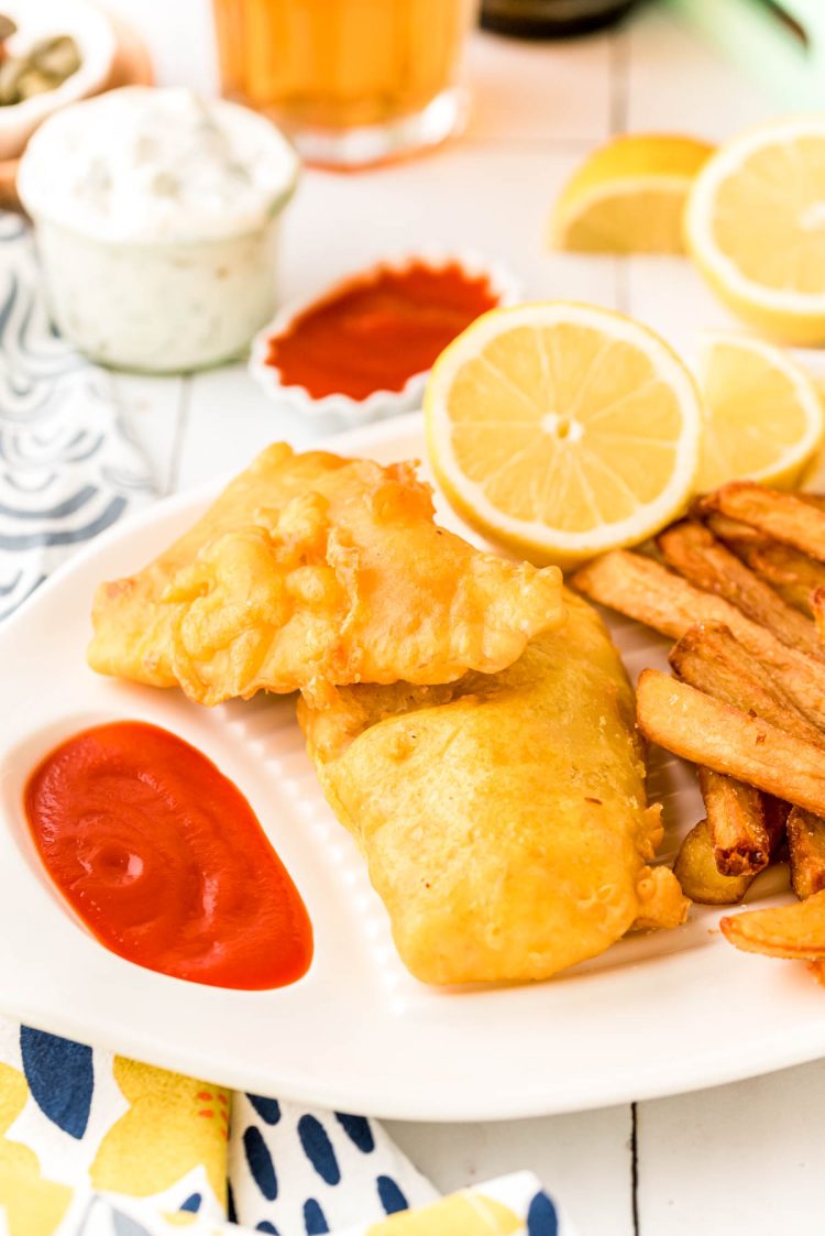 Pieces of fried fish on a white platter with fries and ketchup and lemon.