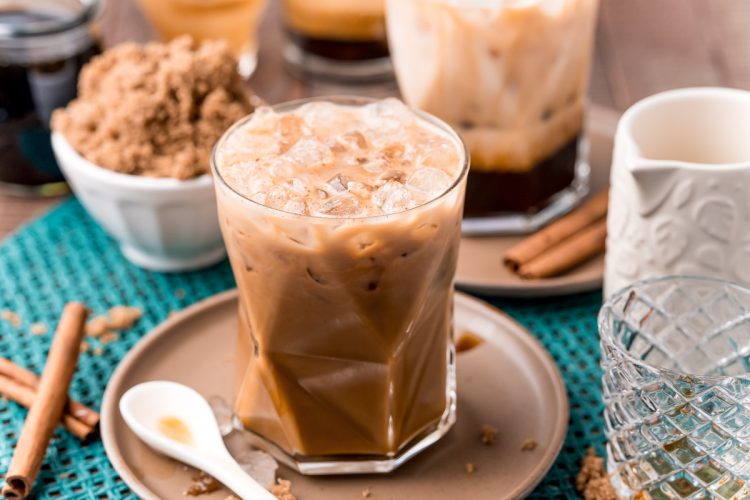 Close up photo of a brown sugar oatmilk shaken espresso on a brown plate with cinnamon sticks and brown sugar.