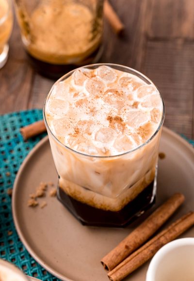 Close up photo of a brown sugar oatmilk shaken espresso on a brown plate with cinnamon sticks and brown sugar.