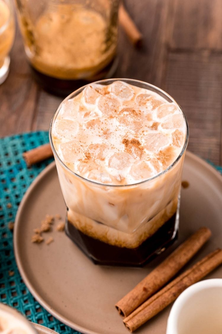 Close up photo of a brown sugar oatmilk shaken espresso on a brown plate with cinnamon sticks and brown sugar.