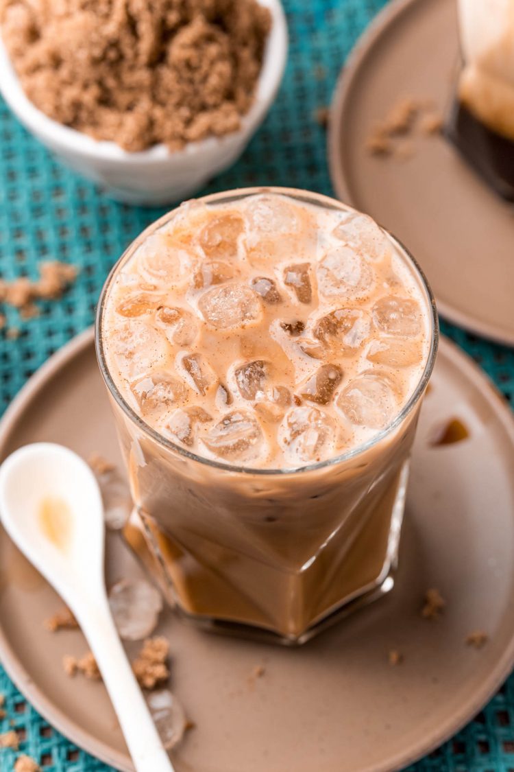 Close up photo of a brown sugar oatmilk shaken espresso on a brown plate with cinnamon sticks and brown sugar.