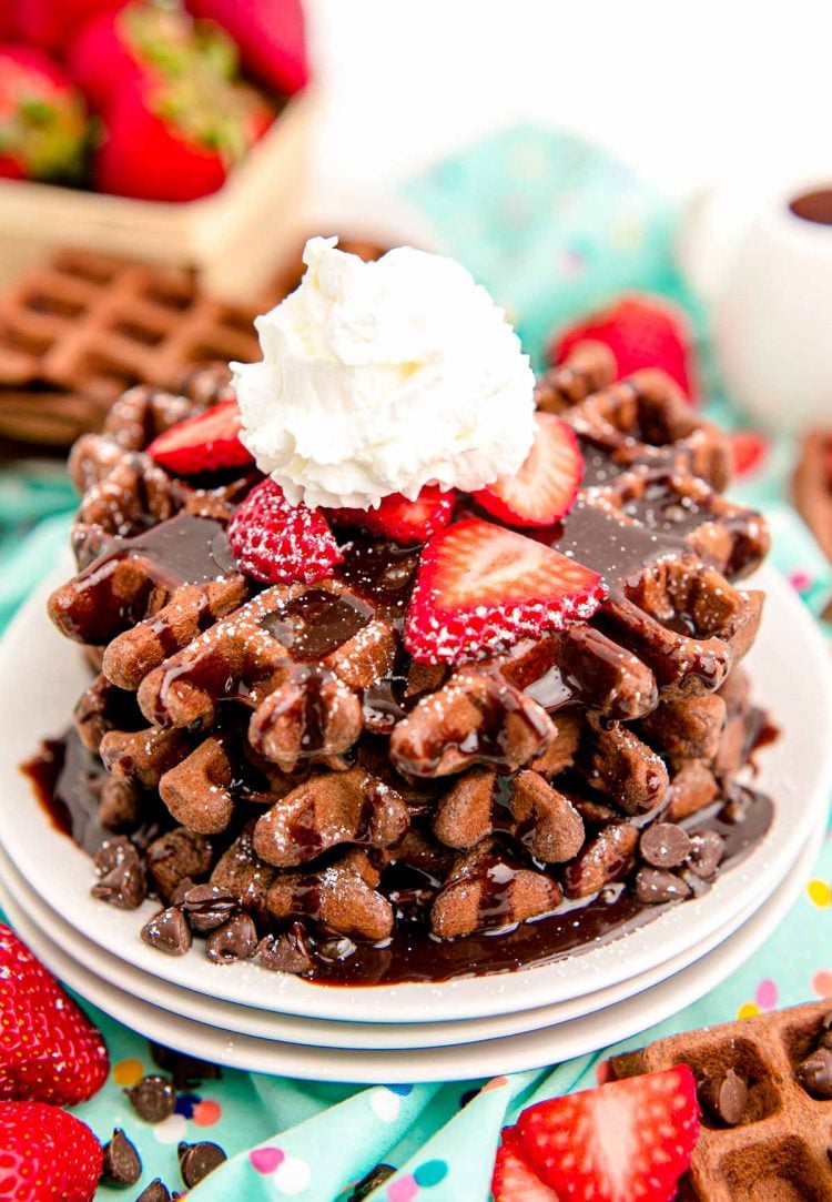 Close up photo of a stack of chocolate waffles topped with chocolate syrup, strawberries, and whipped cream.